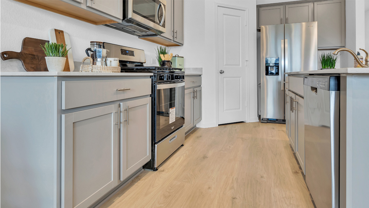 Modern kitchen with light wood flooring, gray cabinets, stainless steel appliances including a fridge and oven. Countertops are decorated with potted plants and kitchen items, creating a clean, fresh feel.