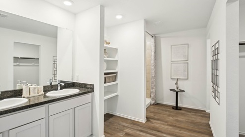 Primary bathroom with dual sinks, walk-in shower, storage shelves, white walls, and vinyl wood flooring.