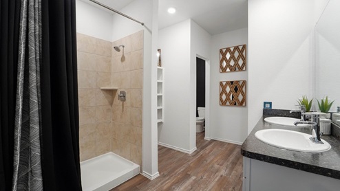 Primary bathroom with dual sinks, walk-in shower, storage shelves, white walls, and vinyl wood flooring.