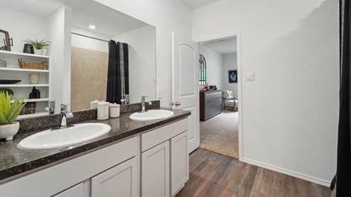 Primary bathroom with dual sinks, walk-in shower, storage shelves, white walls, and vinyl wood flooring.