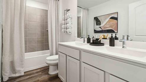 Secondary bathroom with dual sinks, shower and tub combo, white walls, and vinyl wood flooring.