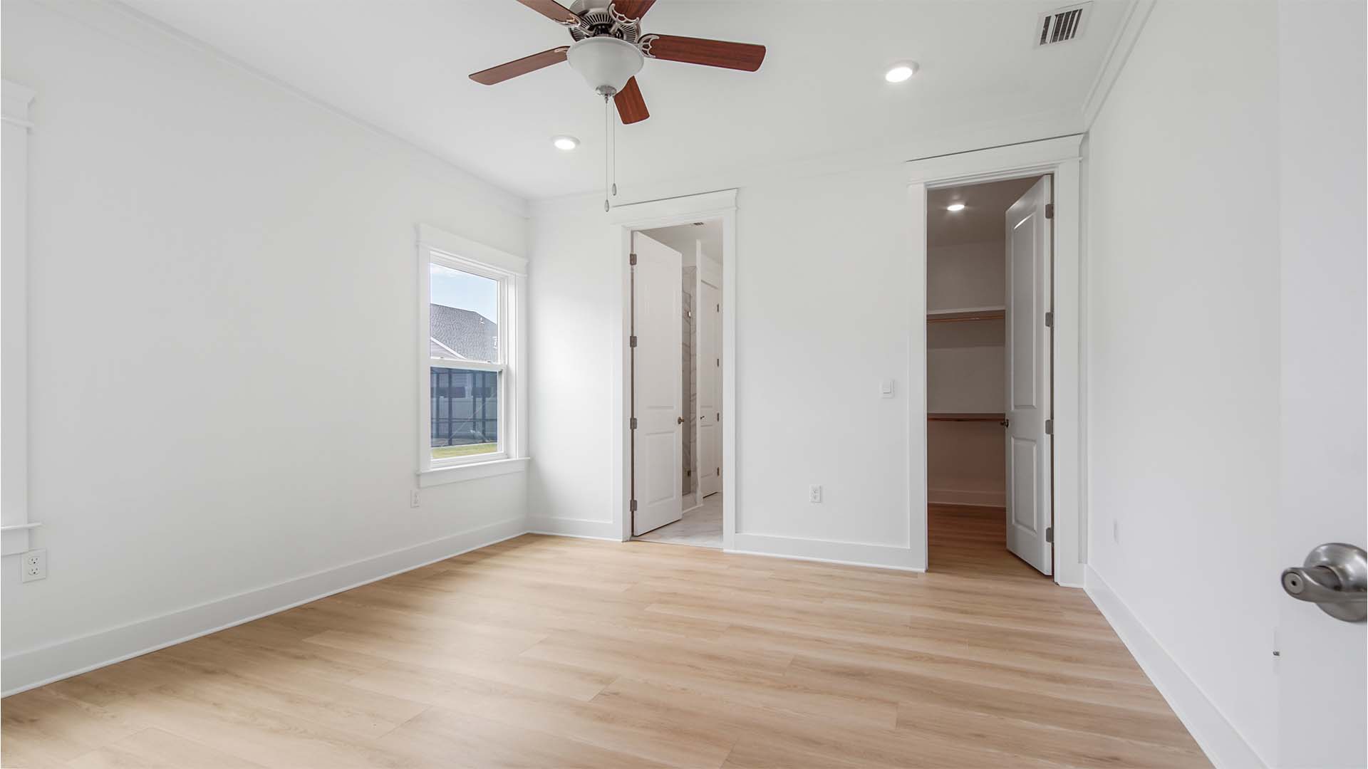 Primary bedroom with EVP flooring and ceiling fan and large windows and walk-in closet with wood shelving and entrance to primary bathroom.
