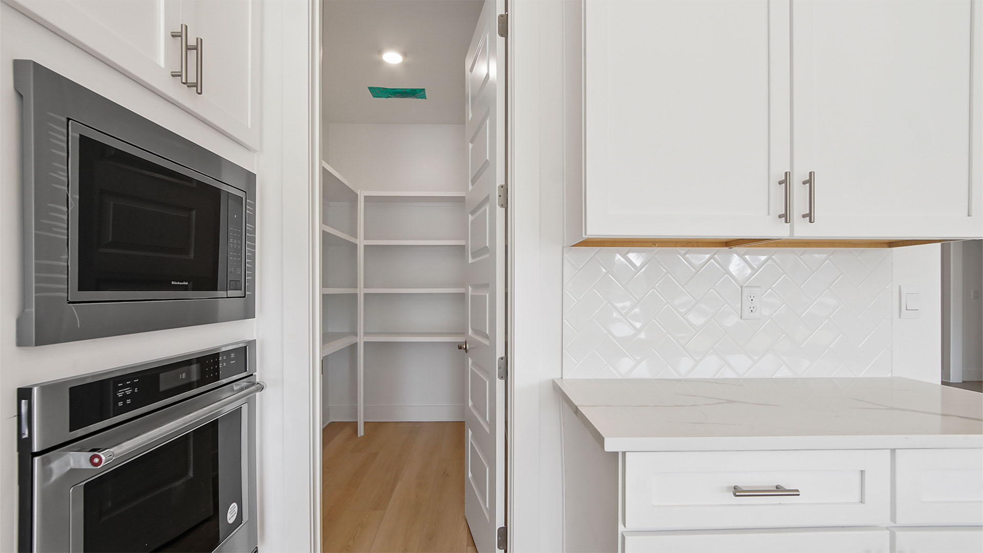 Bright kitchen with white cabinetry and quartz countertops and stainless-steel appliances and entrance to large walk-in pantry with wooden shelving.