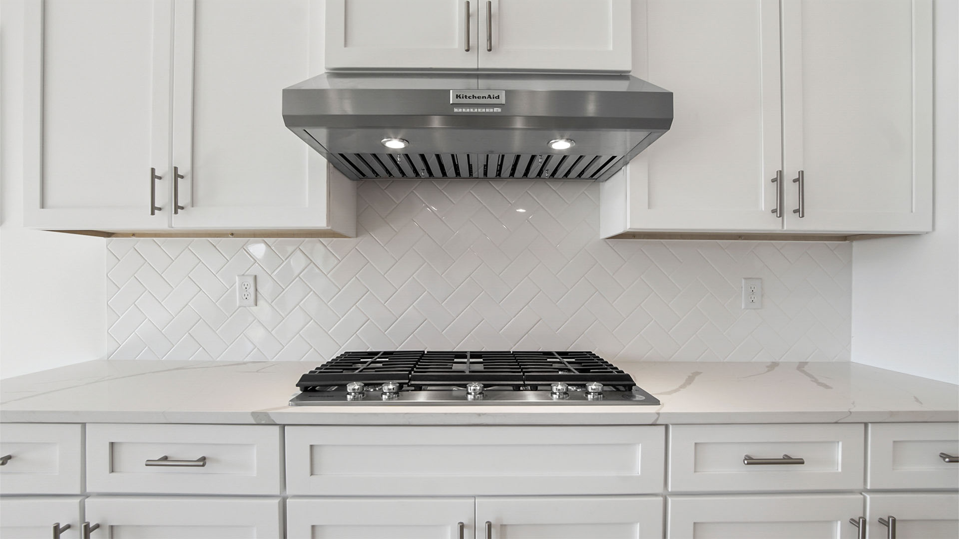Kitchen with stainless-steel appliances and gas-burning stove and range hood and white quartz countertops and herring bone back splash.