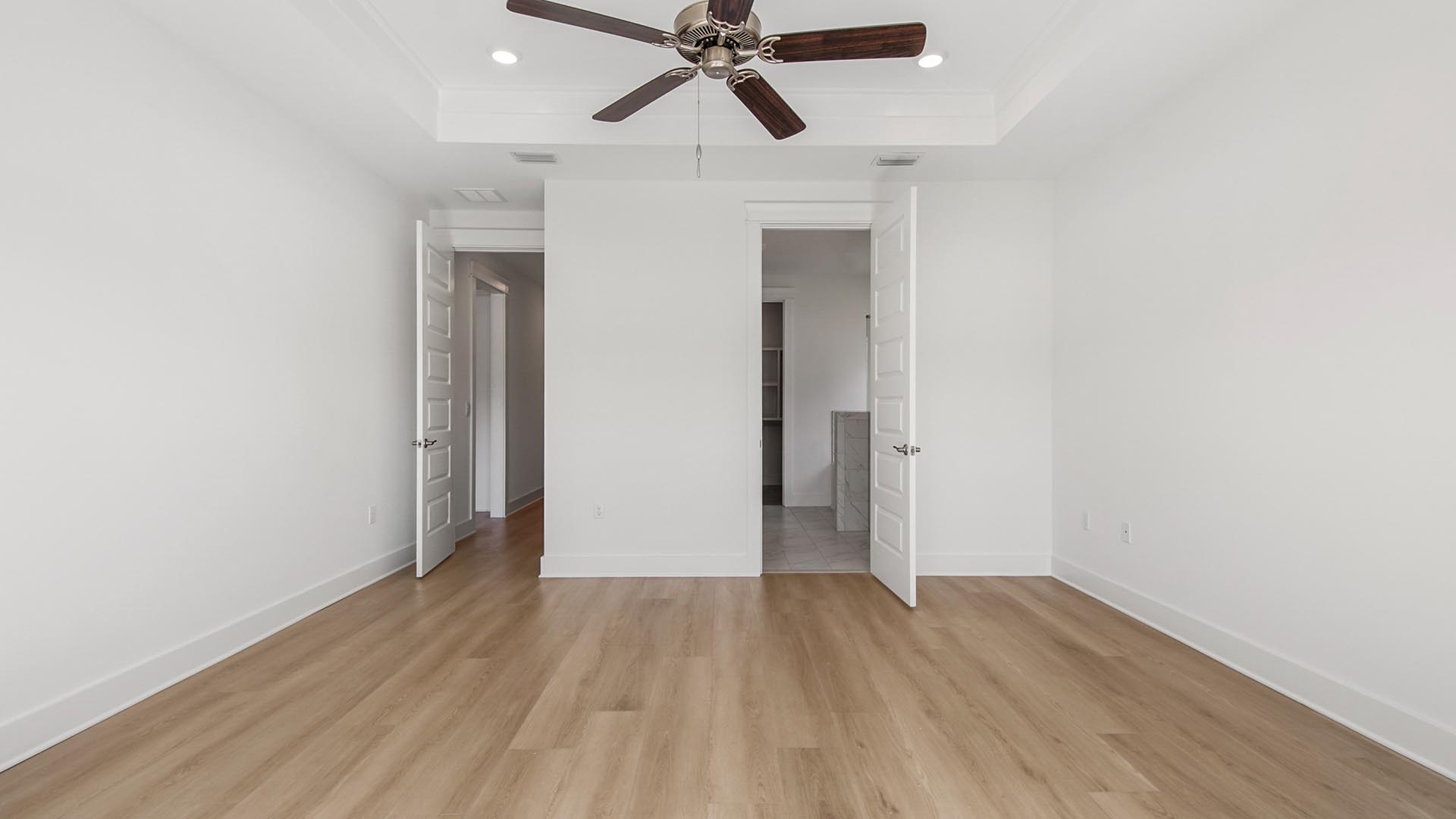 Primary bedroom with EVP flooring and ceiling fan and tray ceiling and door to primary bathroom.