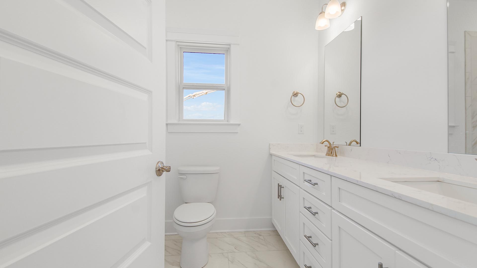 Bathroom three with double vanity with quartz countertops and white cabinetry and two mirrors and marbled tile flooring and window above toilet.
