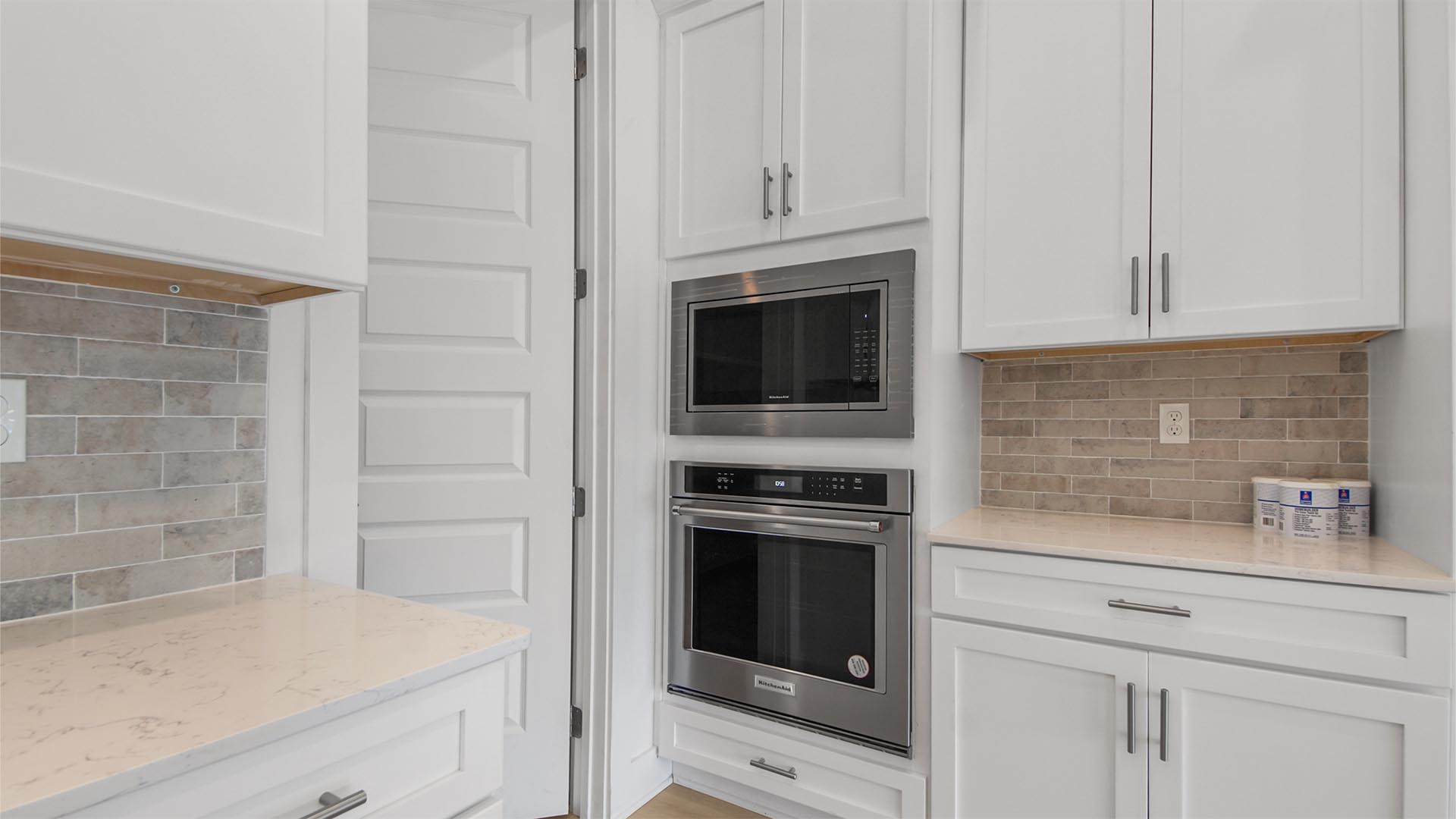 Stainless-steel oven and microwave and tall white kitchen cabinets and Herringbone backsplash and tall door to walk-in pantry.