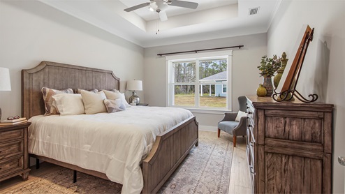 Breakfast Point Bluewater plan primary bedroom with EVP flooring and a large window and a ceiling fan and tray ceilings.