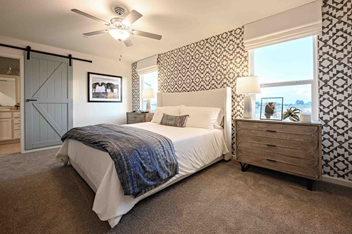 Interior photo of primary suite with bed, end tables, dresser, and large windows.