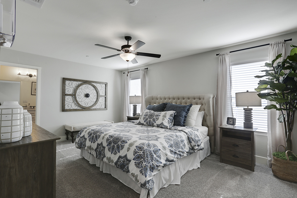Interior photo of primary suite with bed, end tables, dresser, and large windows.