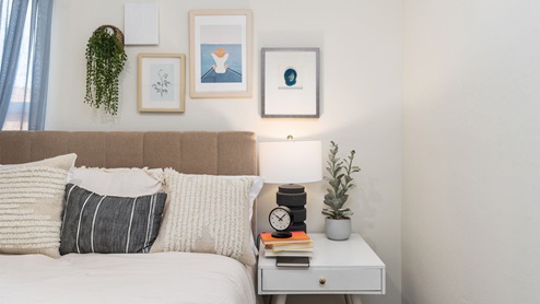 A close up picture of a bed with decorative pillows, an end table with decor, and art on the wall