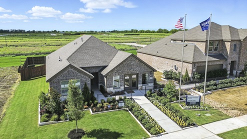 Front exterior of a one story model home with a brick exterior and luscious green landscaping