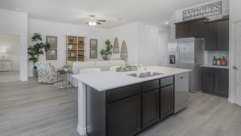 Kitchen with island surrounded by brown cabinets with hard surface countertops and stainless steel appliances and views into the living room