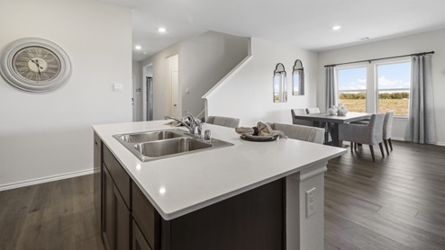 Kitchen island surrounded by white counters, brown cabinets and stainless steel appliances and views into the living room