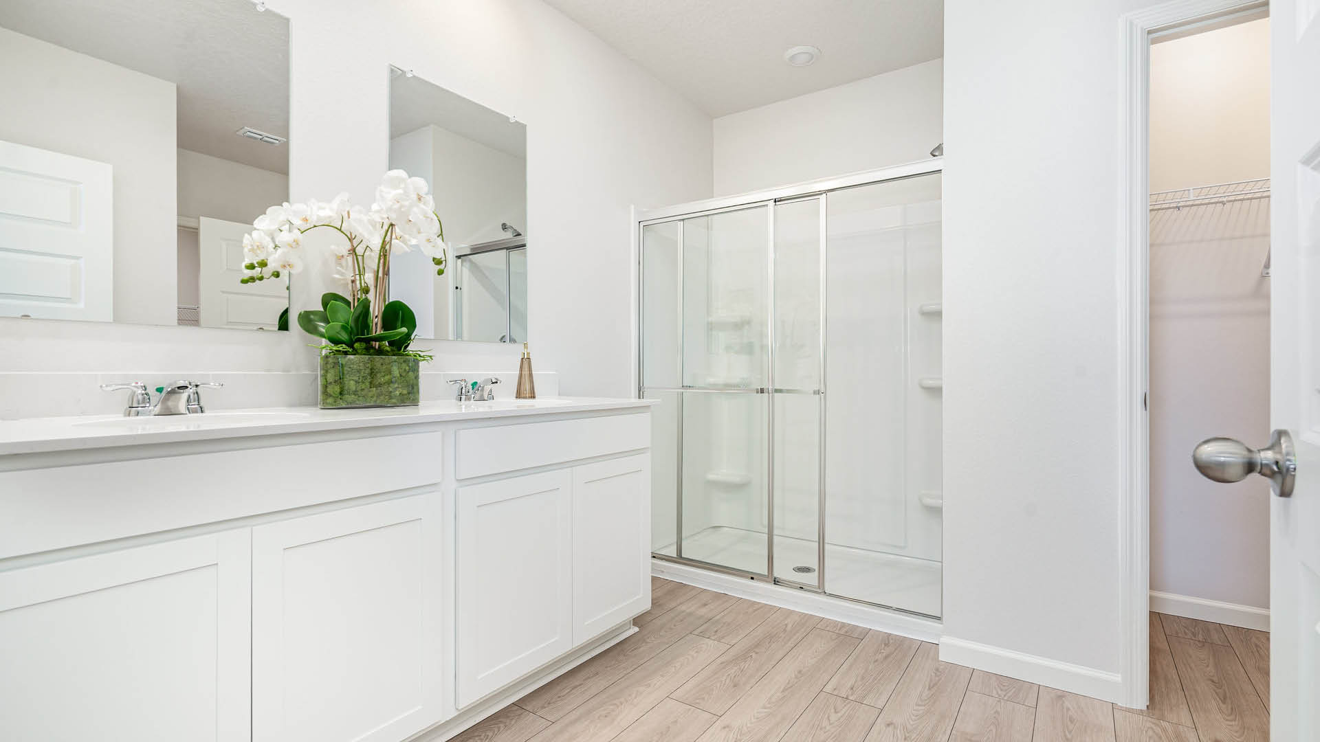 Spacious white bathroom with dual sinks on the left, a glass-door shower on the right, and a potted orchid adds elegance on the counter. Bright, serene feel.