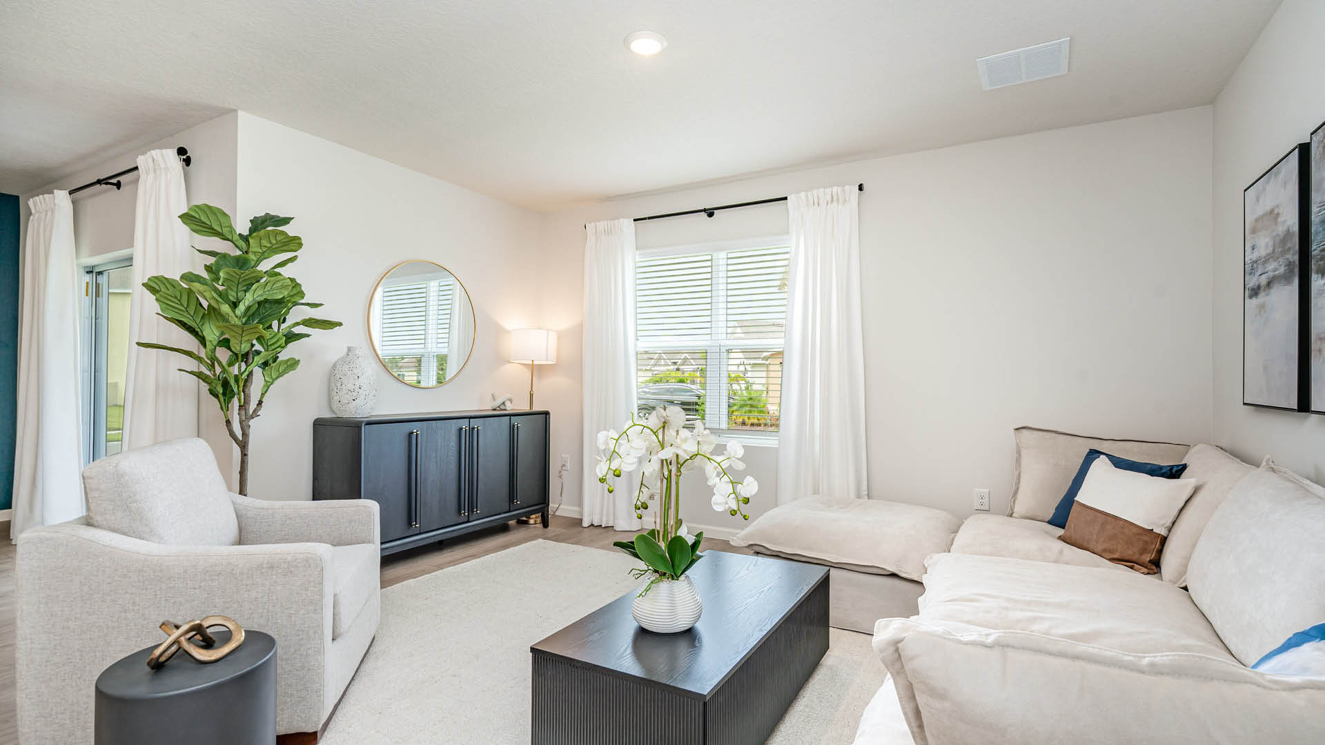 Bright and stylish living room featuring a beige sectional sofa, a coffee table with a plant centerpiece, and large windows with sheer curtains.
