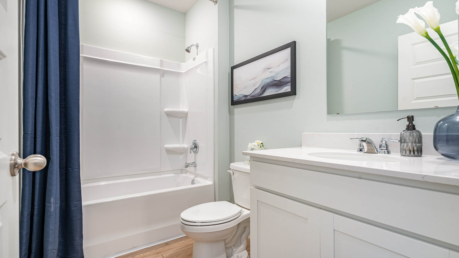A modern bathroom featuring a white bathtub with a dark blue curtain, white vanity with twin faucets, decorative vase with flowers, and an abstract wall art.