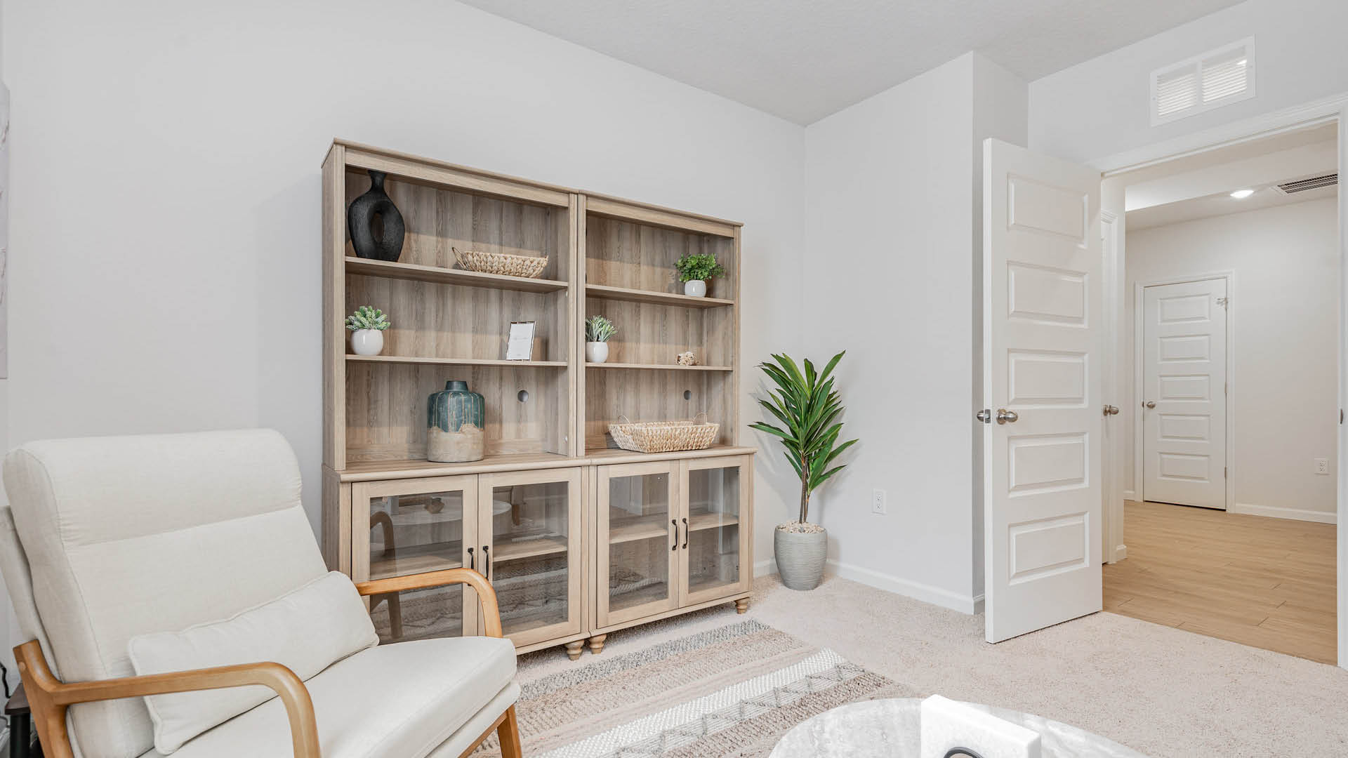 A cozy room with beige walls features a light wooden bookshelf, accented by plants and decor. A plush chair and a closed door enhance the inviting atmosphere.