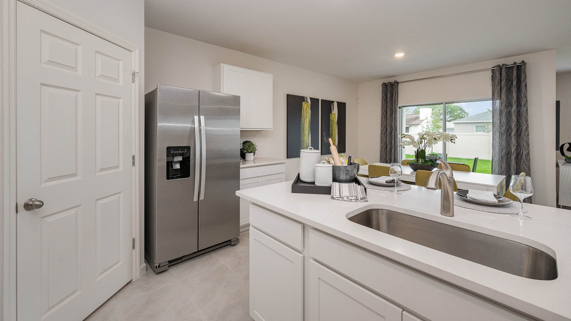 Modern kitchen featuring stainless steel refrigerator, white cabinetry, and a dining area with elegant table settings and large windows.