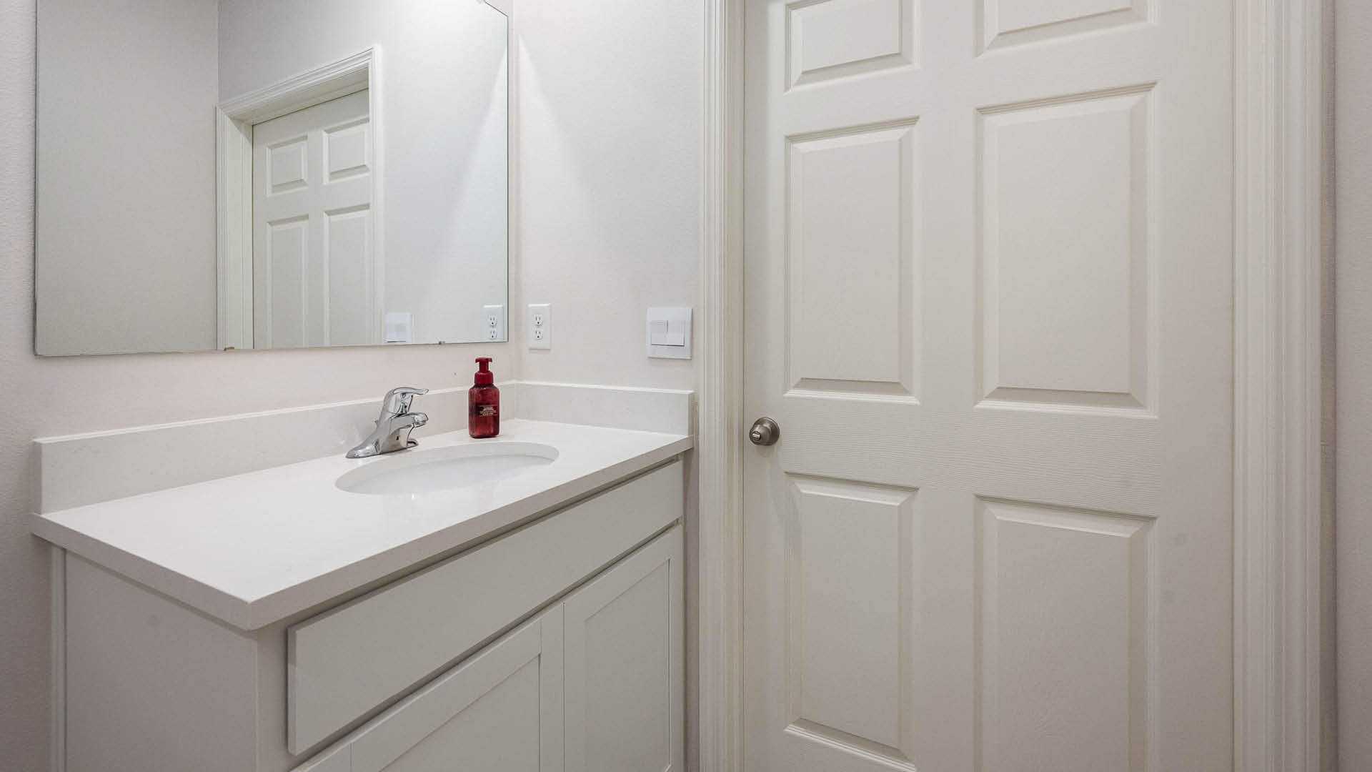 A modern bathroom featuring a white vanity with a sink, mirror, and a red soap dispenser, beside a closed door.
