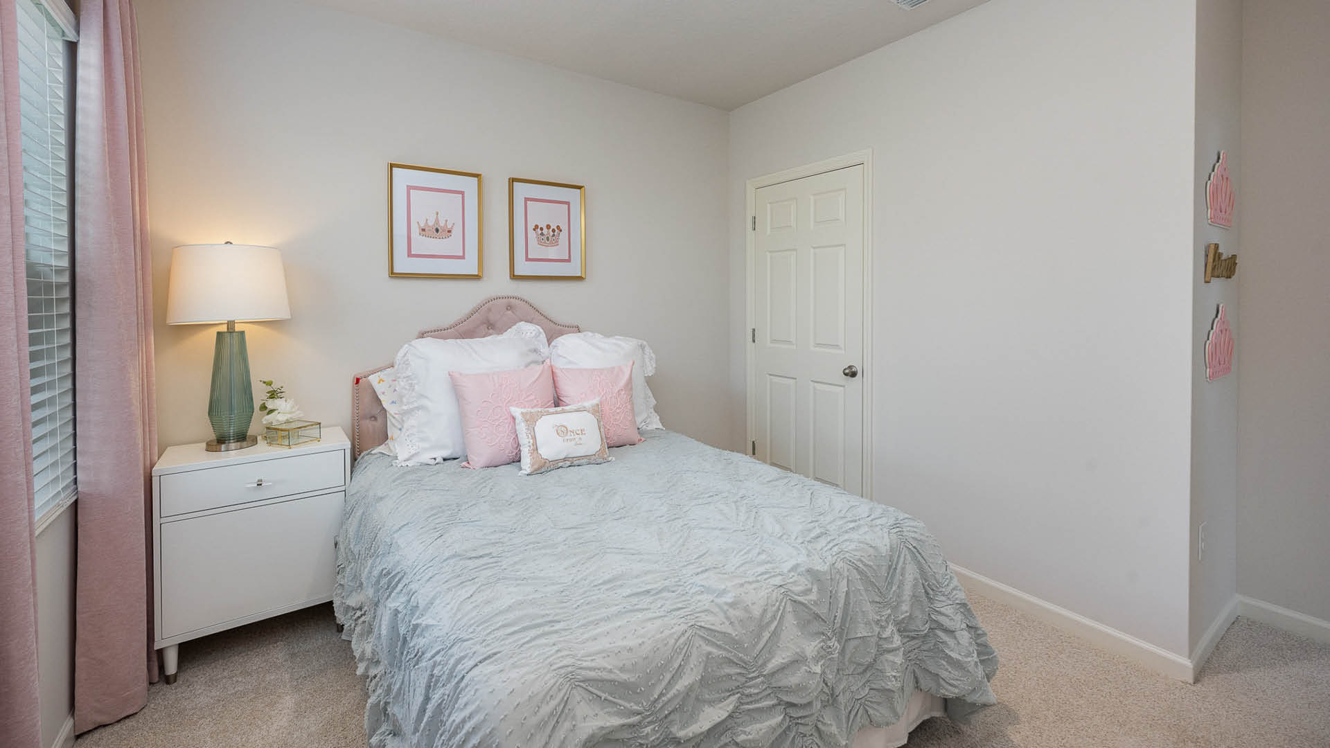 Cozy bedroom featuring a light blue bedspread, pink accents, a stylish lamp, and framed crown art on the wall, with soft carpet and curtains.
