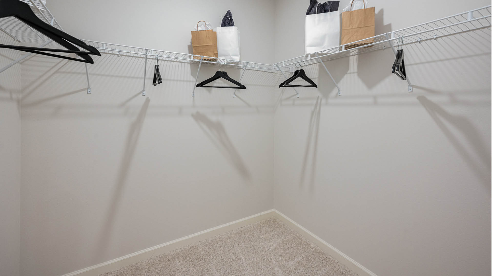 A spacious, empty closet with wire shelving, black hangers, and gift bags, all against a light gray wall and carpeted floor.