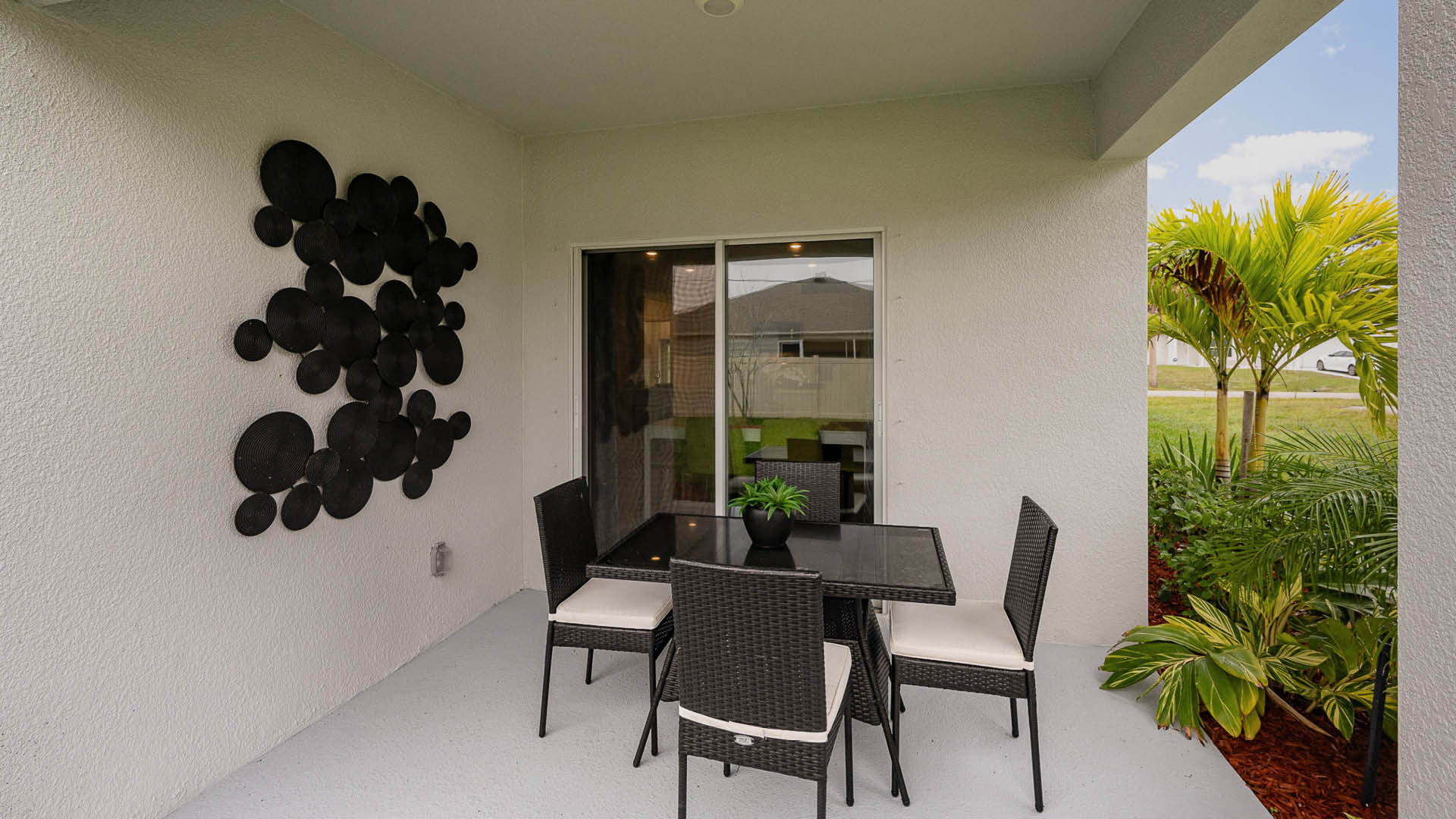 A modern patio with a glass-top table, four wicker chairs, and decorative wall art, surrounded by greenery visible through sliding doors.