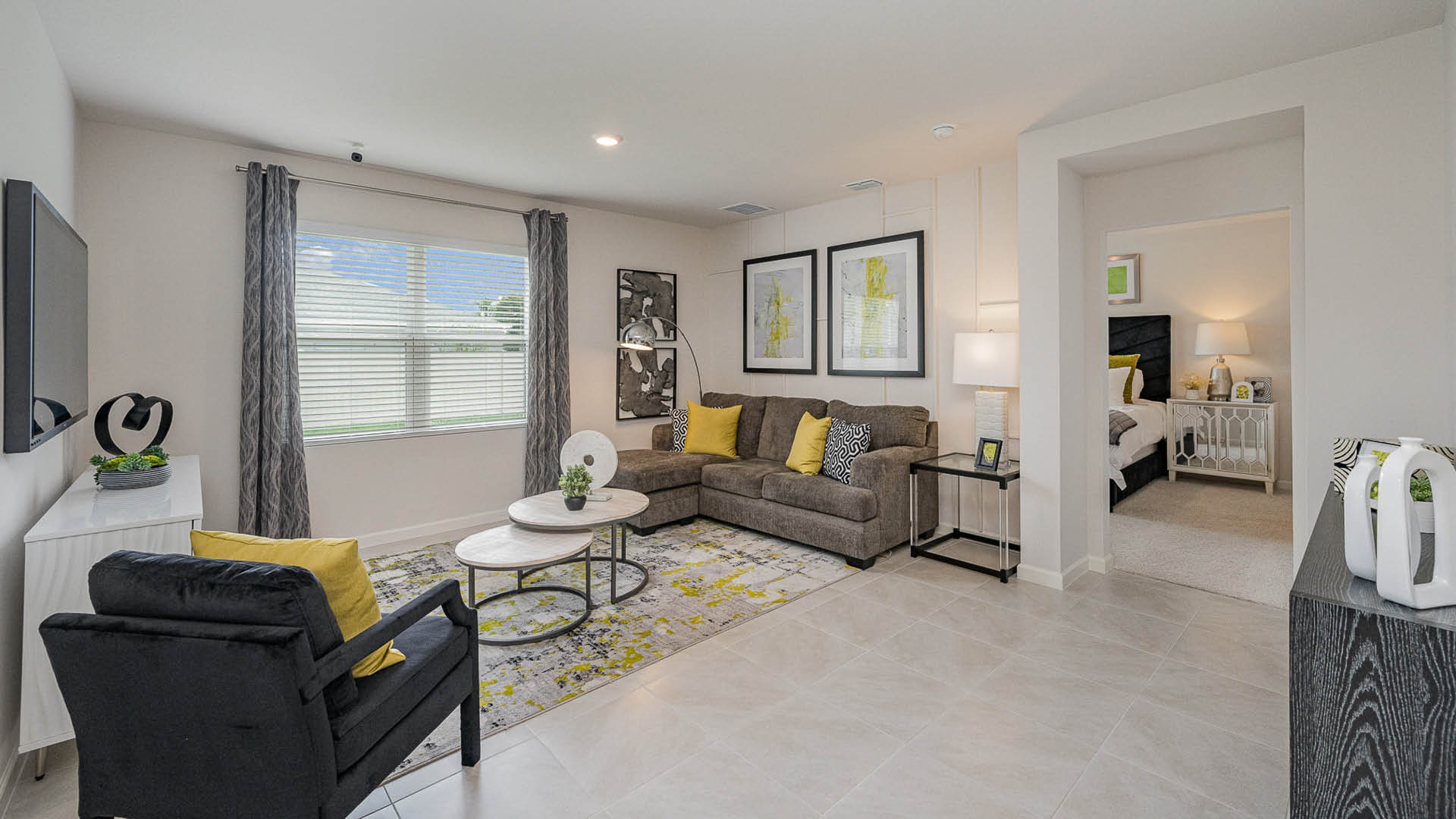Modern living room featuring a gray sofa with yellow accents, a round coffee table, and decorative art, leading to a cozy bedroom space.