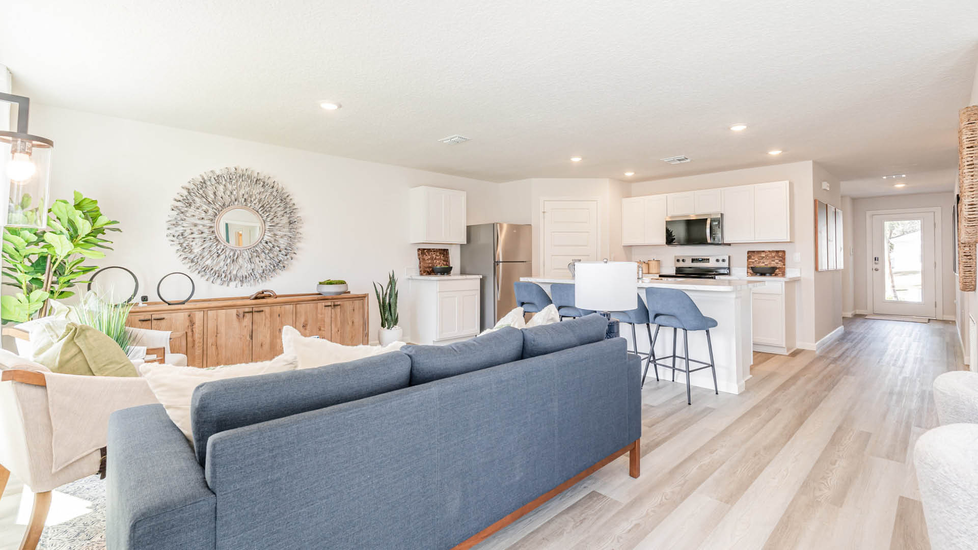 Bright, modern living room with a blue couch, wood accents, and a kitchen area featuring white cabinets and stainless appliances.