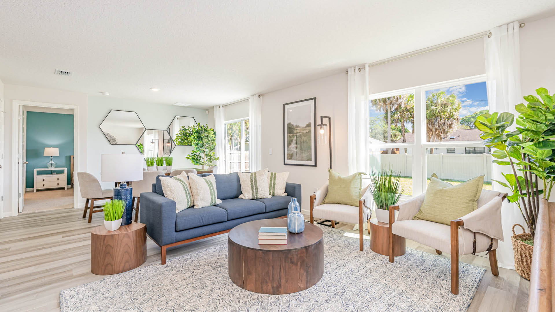 Bright and airy living room featuring a blue sofa, two armchairs, a round coffee table, and plants, with large windows and modern decor.