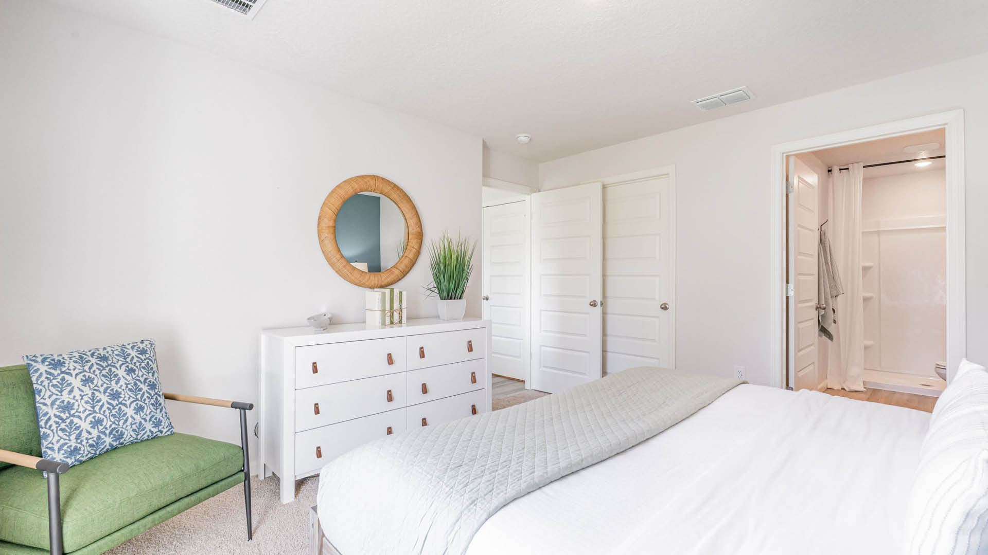 The image features a clean and organized bedroom with a large bed occupying the majority of the space. The bed is positioned against the wall, with a mirror hanging above it. The room also contains a dresser and a potted plant, adding a touch of greenery to the space.   In addition to the main furniture, there are two chairs in the room, one located near the left side of the bed and the other closer to the center. The room appears to be well-maintained and ready for use.