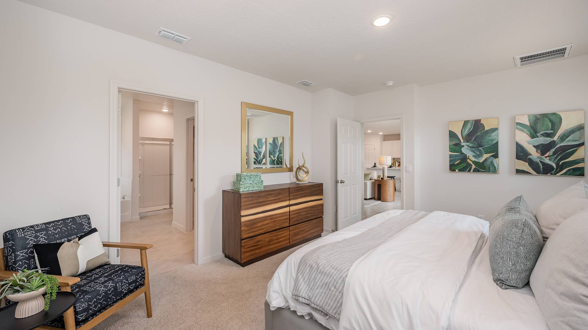 Bright and cozy bedroom featuring a plush bed, modern dresser, lounging chair, and green plant decor, with a bathroom entrance visible.