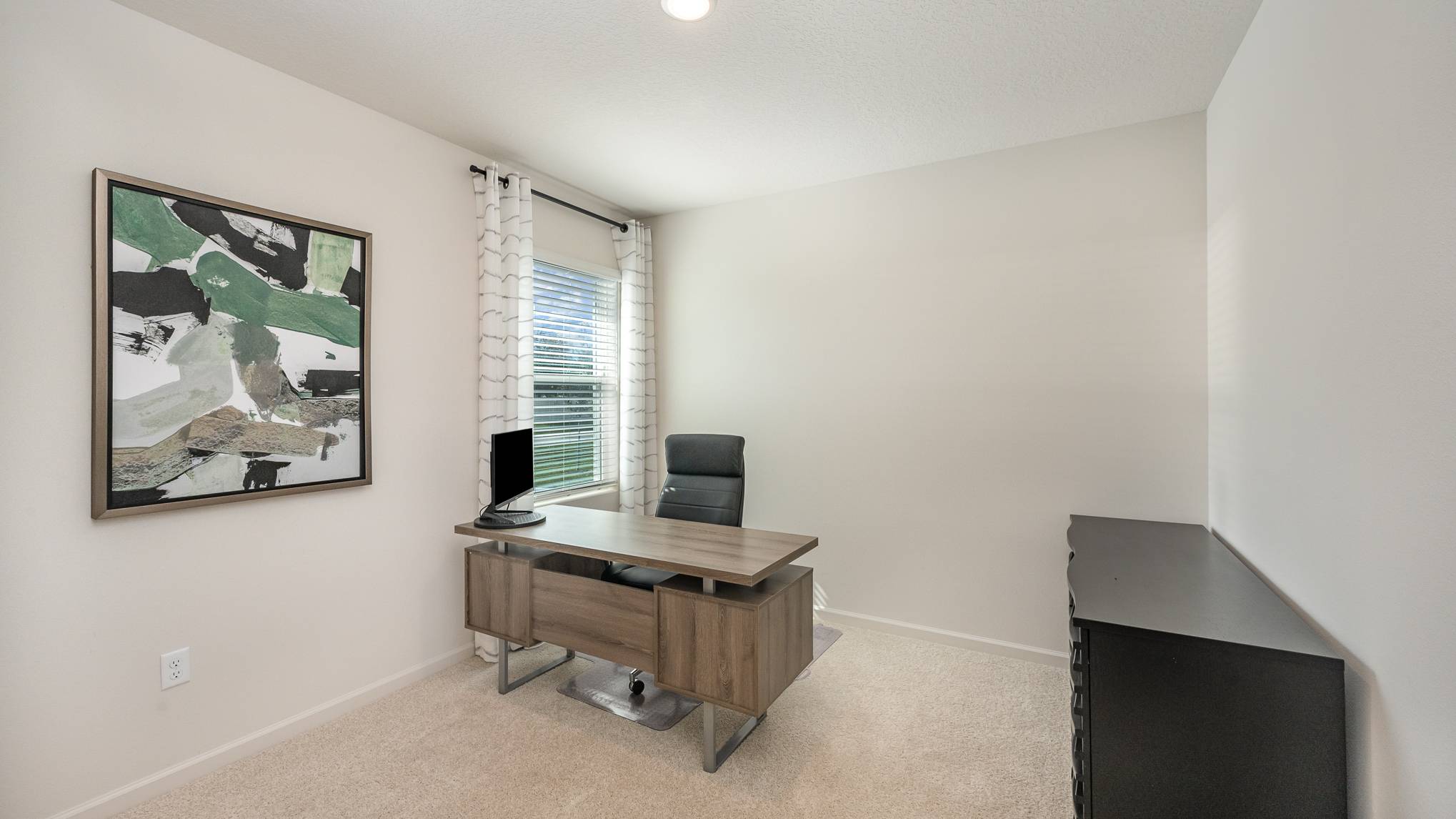 A modern home office featuring a wooden desk with a computer, a black ergonomic chair, and a window with light curtains.