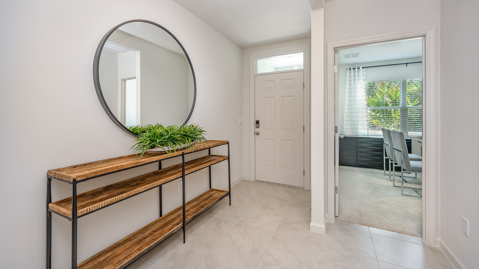 Bright entryway featuring a round mirror, wooden shelf with greenery, and an open door leading to a well-lit room with modern chairs.