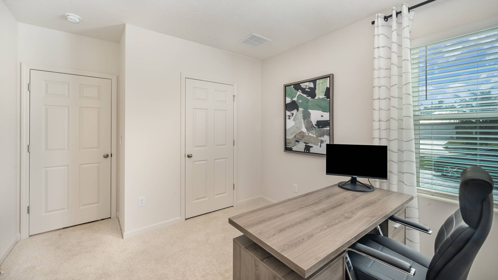 Modern home office with beige carpet, a wooden desk, a black chair, and a large window with white curtains. Two doors are visible.