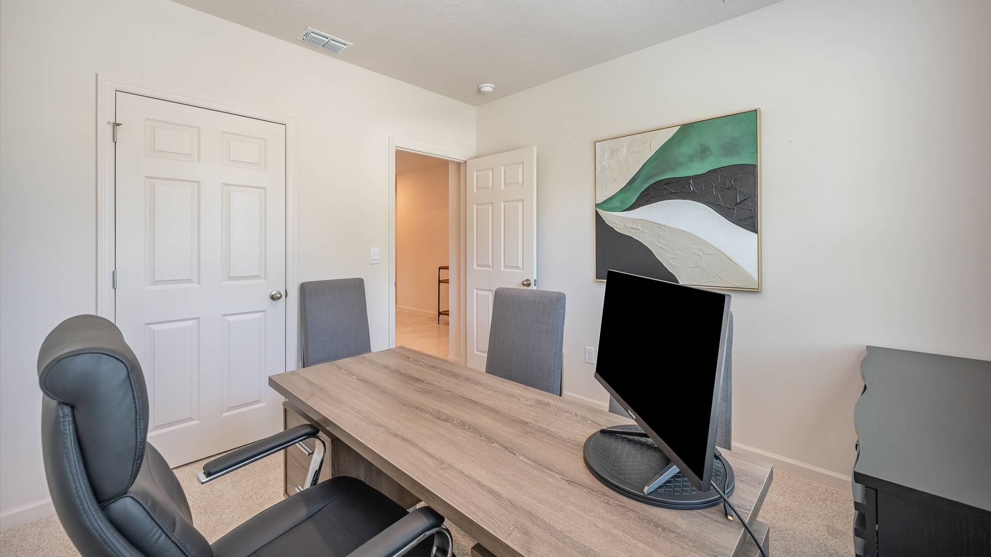 Modern office interior with a wooden desk, two upholstered chairs, a blank monitor, and abstract artwork on the wall.