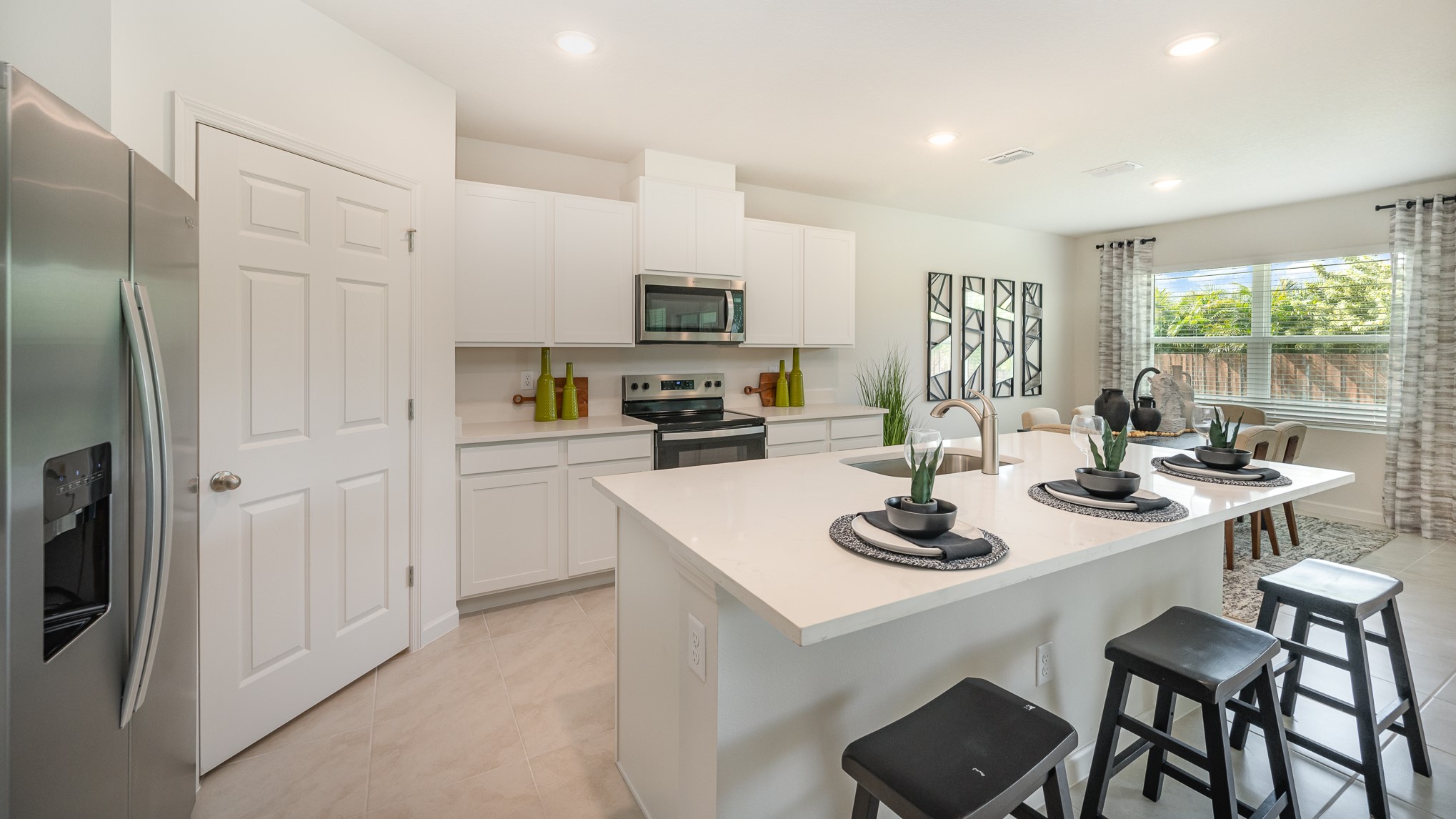Modern kitchen with white cabinetry, stainless steel appliances, an island with black stools, and decorative plants and table settings.