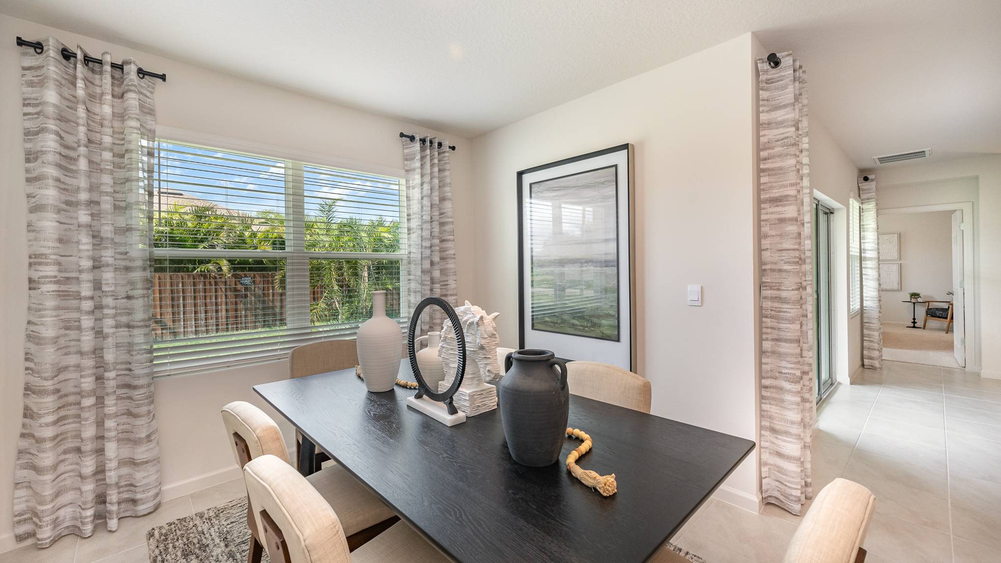 Bright dining area with a black table, decorative vases, and art, framed by sheer curtains, overlooking a green backyard.