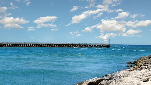 Sunny seaside view featuring a long pier stretching into calm blue waters, with boats and scattered clouds above.