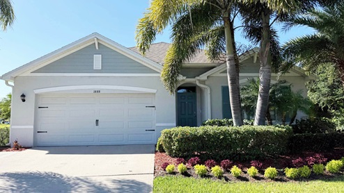 Single-story home with a grey and white facade, surrounded by lush greenery and colorful landscaping, under a clear blue sky.