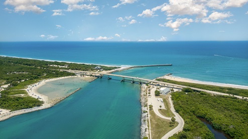 Aerial view of a coastal area featuring a bridge, sandy beach, lush greenery, and the ocean under a clear blue sky with scattered clouds.