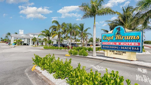 Sign for Capt Hiram's Resort with palm trees and a clear blue sky, advertising river dining, beach bar, and wine tasting event.