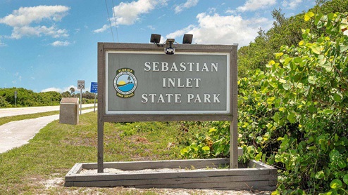 Sign for Sebastian Inlet State Park, featuring the park's name, logo, and nearby speed limit sign against a blue sky and green foliage.