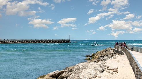 A scenic view of a pier extending into the turquoise ocean, with people enjoying the day and boats passing by under a bright sky.