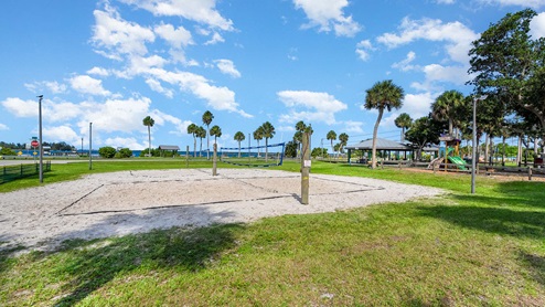 A sandy volleyball court in a grassy park surrounded by palm trees, with picnic pavilions and a playground in the background.