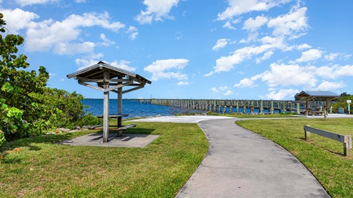 A scenic path leads to the ocean with a wooden gazebo and a long pier in the background against a bright blue sky and fluffy clouds.