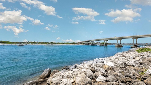Scenic view of a calm blue river, a bridge arching overhead, and rocky shoreline under a bright sky with fluffy clouds.