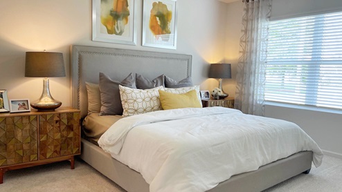 Cozy bedroom with a gray upholstered bed, decorative pillows, stylish lamps, and a wooden nightstand beside a window with blinds.