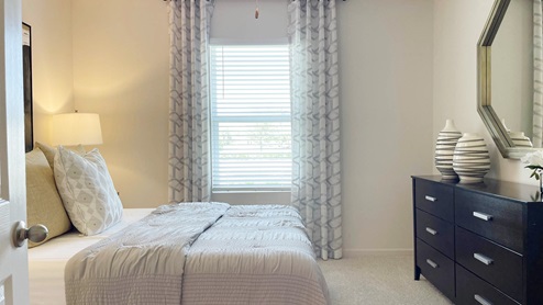 Cozy bedroom featuring a bed with decorative pillows, a window with blinds, and a dresser adorned with vases and a mirror.