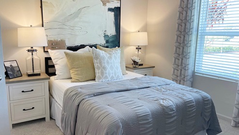 Cozy bedroom featuring a twin bed with gray bedding, decorative pillows, bedside lamps, and abstract wall art. Natural light through windows.